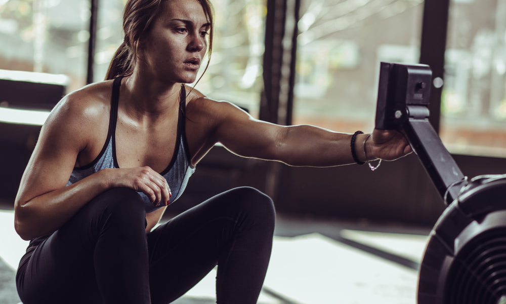 A woman in workout clothes sits on a rowing machine and reaches out to tocuh the screen on the attached control montior.