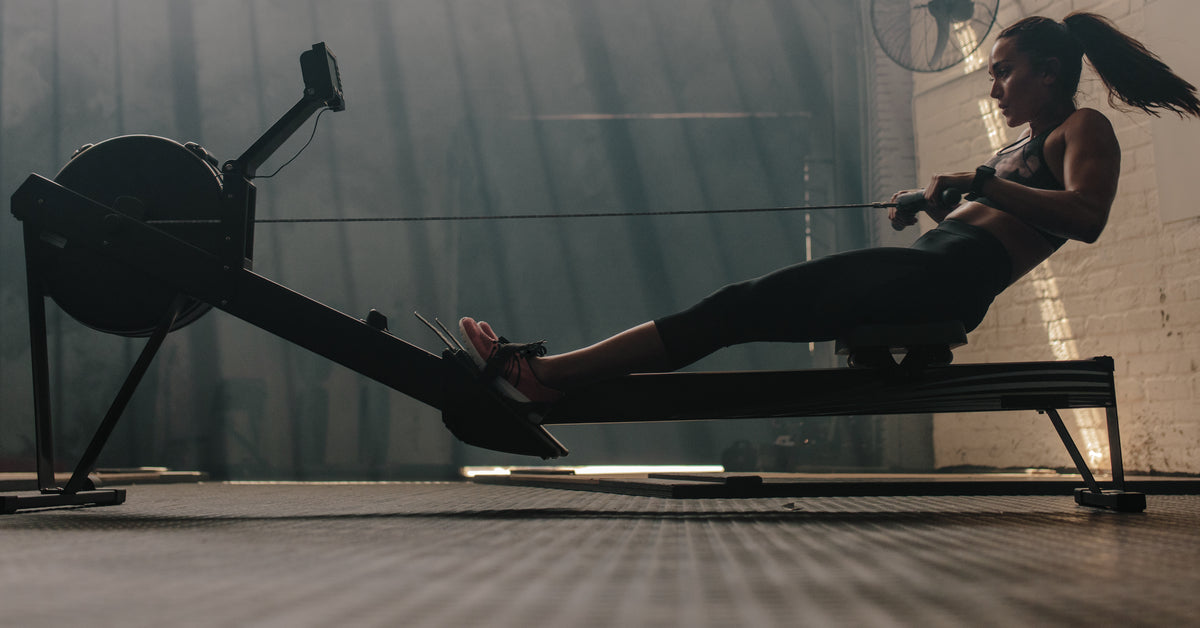A woman in a gym pulls the handle of a rowing machine. Small streams of light enter high windows of the dimly lit gym.