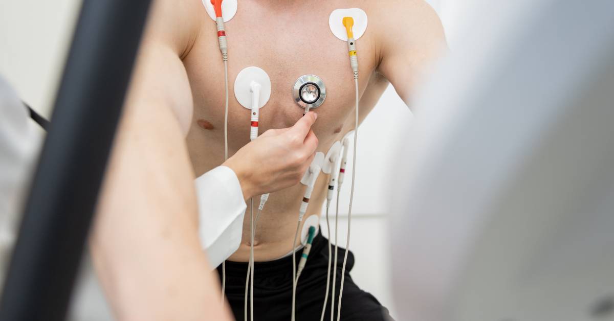 A hand attaches electrodes to various parts of a man's chest as he prepares for a physical fitness test on an exercise bike.