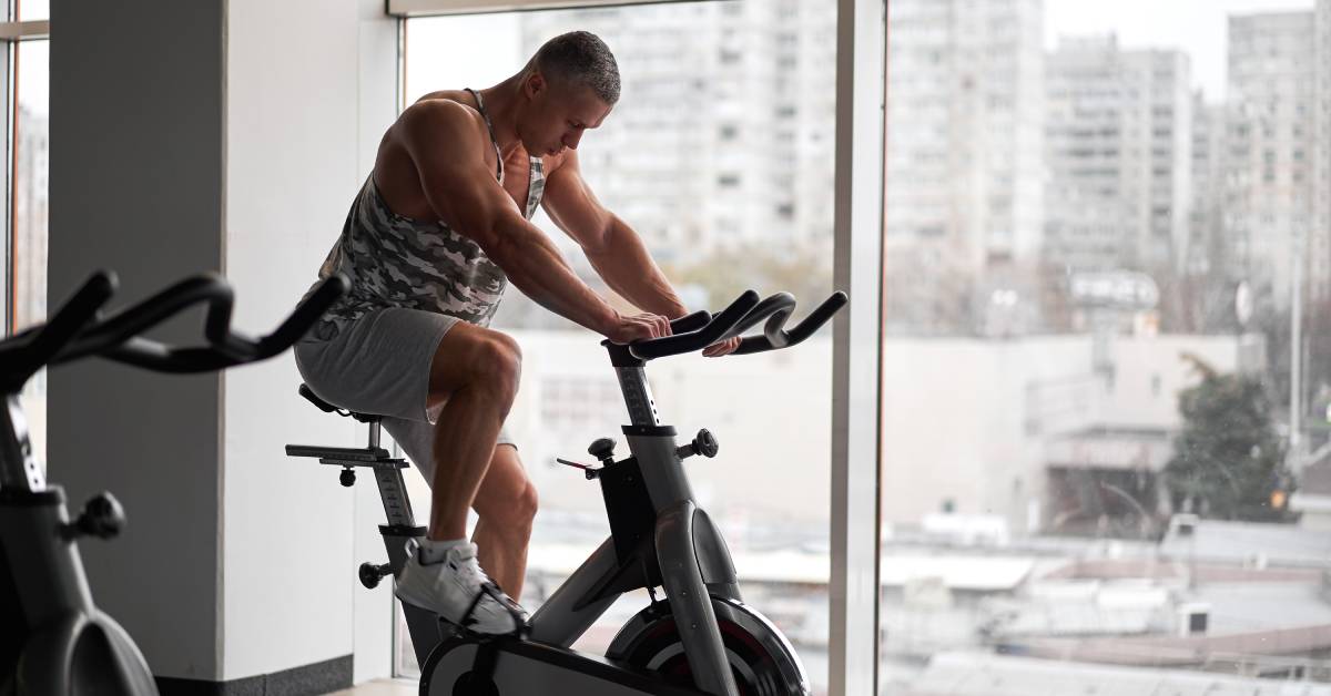 A man in a gray tank top pedals a stationary bike in a gym as the light from the large window next to him casts shadows.