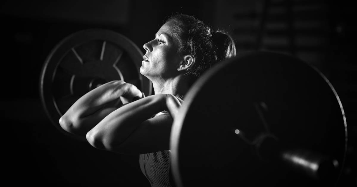 A woman in a black and white setting holding a barbell with large plates on both ends and her wrists pointed outward.