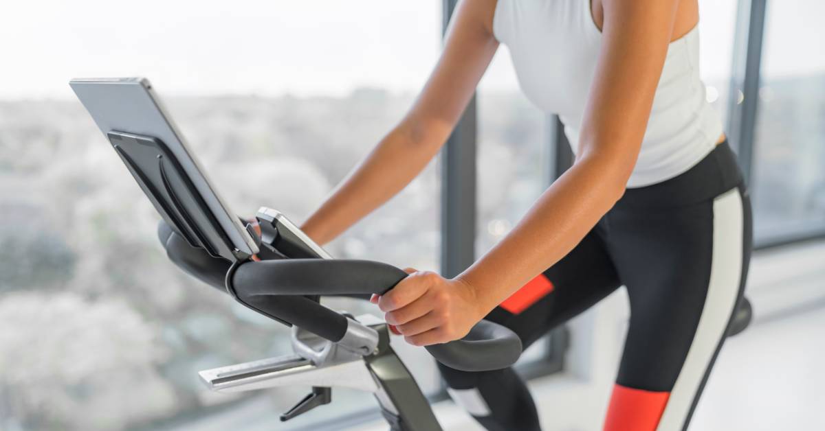 A women in a white shirt and orange, black, and white fitness pants riding an indoor bike next to a large window.