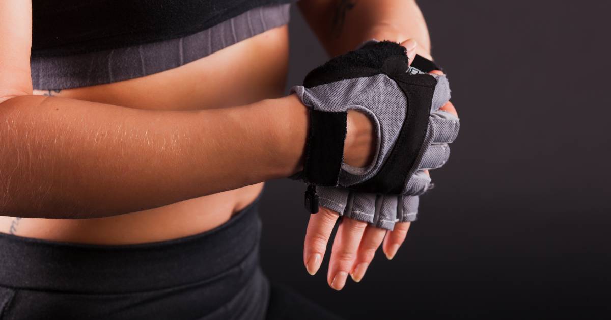 A woman in a black top and black pants secures black and gray fingerless fitness gloves onto her hands.