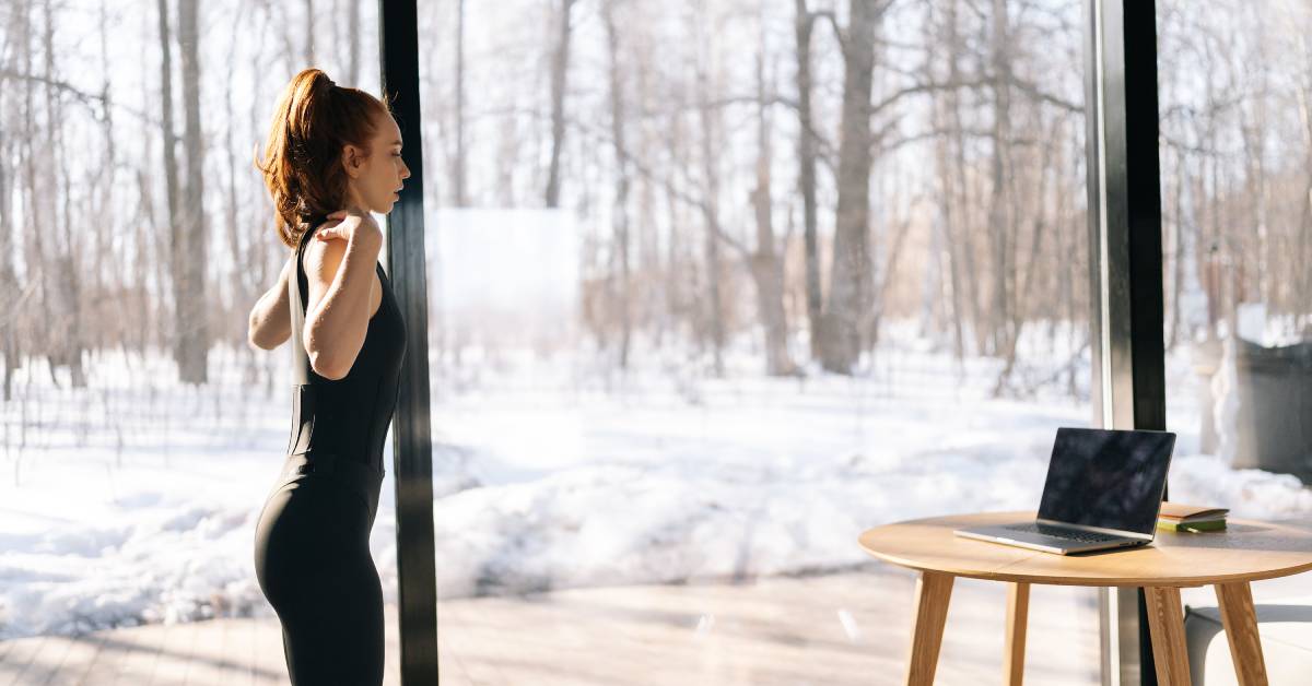 A woman in black workout clothes standing up and looking at a laptop with her arms out and hands touching her shoulders.
